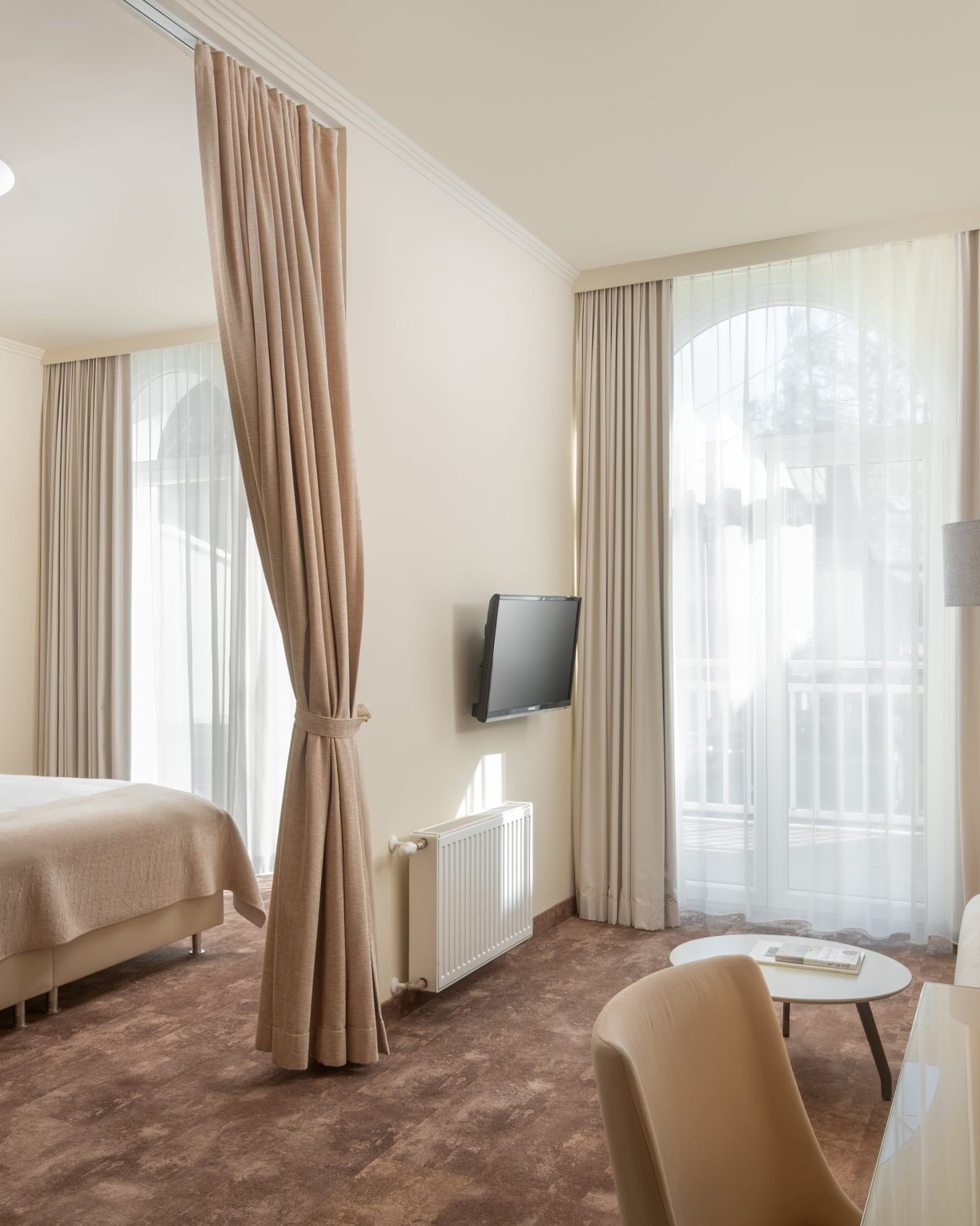 Helles Standard Plus Doppelzimmer im Hotel Post Wrann in Velden am Wörthersee, Kärnten. Das geräumige Zimmer zeigt ein großes Doppelbett mit beiger Bettwäsche, eine gemütliche Sitzecke mit Sofa, Schreibtisch und TV. Durch die großen Fenster mit Vorhängen fällt viel Tageslicht, das eine freundliche, entspannte Atmosphäre schafft.