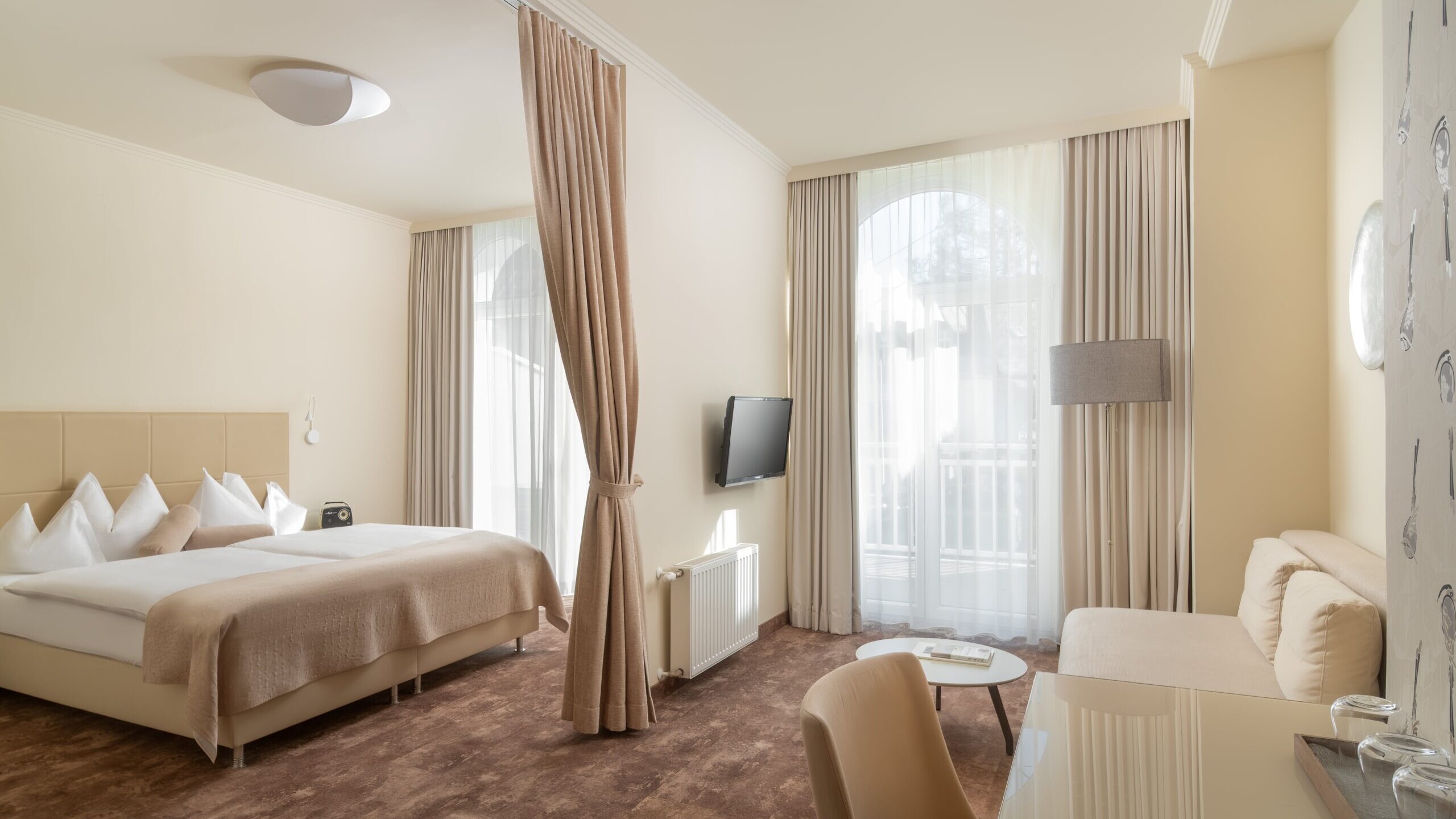 Helles Standard Plus Doppelzimmer im Hotel Post Wrann in Velden am Wörthersee, Kärnten. Das geräumige Zimmer zeigt ein großes Doppelbett mit beiger Bettwäsche, eine gemütliche Sitzecke mit Sofa, Schreibtisch und TV. Durch die großen Fenster mit Vorhängen fällt viel Tageslicht, das eine freundliche, entspannte Atmosphäre schafft.