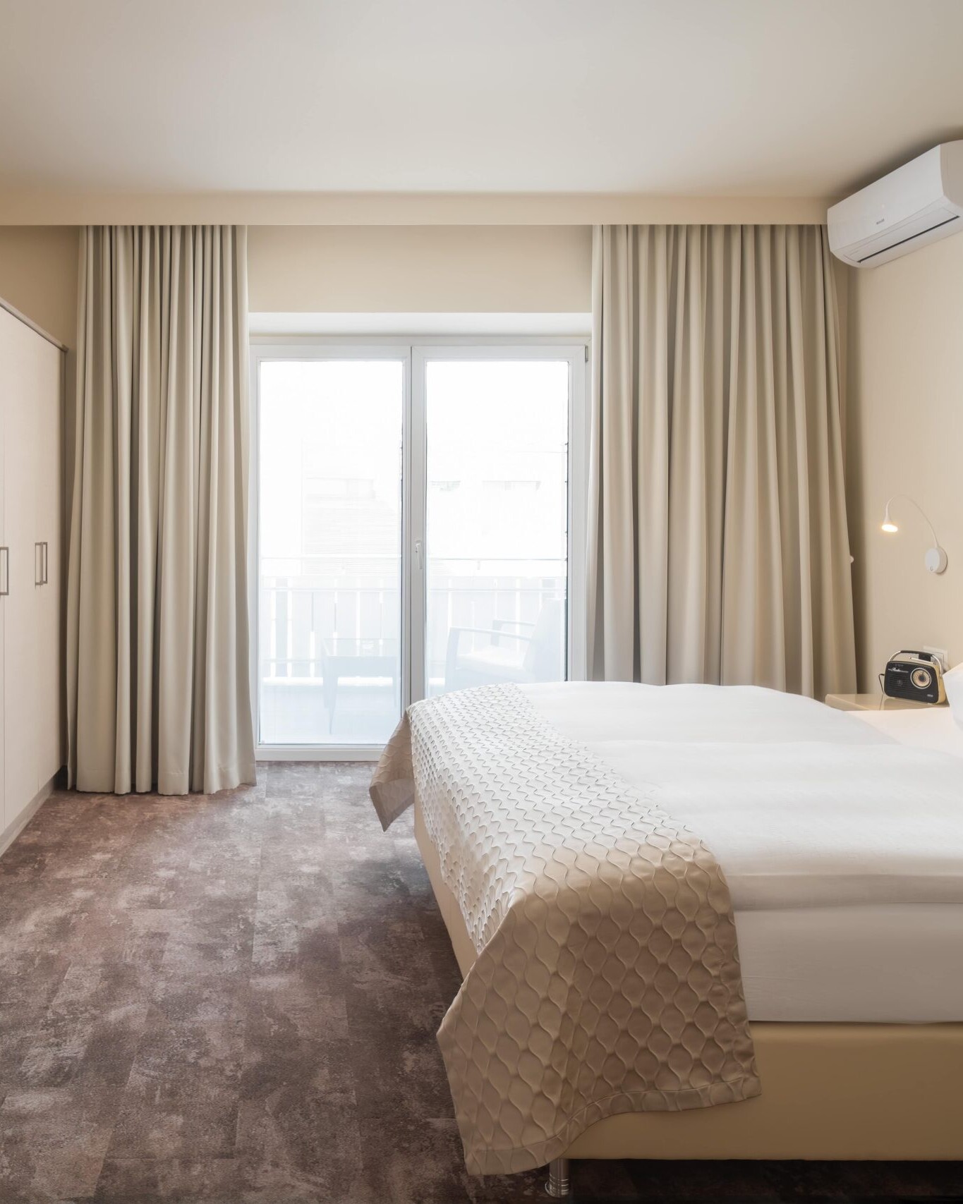 Helles Standard-Doppelzimmer im Hotel Post Wrann in Velden am Wörthersee, Kärnten. Das Zimmer zeigt ein großes Doppelbett mit beiger Bettwäsche, einen Schreibtisch mit Lampe, einen Fernseher und Zugang zu einem Balkon mit Vorhängen. Moderne, gemütliche Atmosphäre für erholsame Nächte.