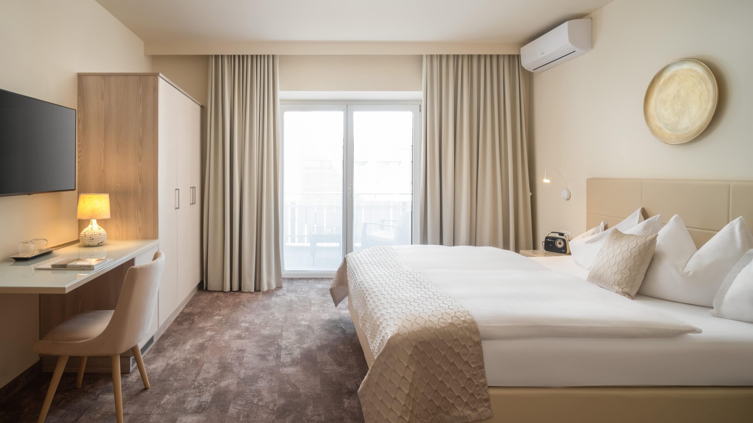 Helles Standard-Doppelzimmer im Hotel Post Wrann in Velden am Wörthersee, Kärnten. Das Zimmer zeigt ein großes Doppelbett mit beiger Bettwäsche, einen Schreibtisch mit Lampe, einen Fernseher und Zugang zu einem Balkon mit Vorhängen. Moderne, gemütliche Atmosphäre für erholsame Nächte.